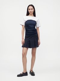 Femme debout portant un haut blanc à manches courtes avec un empiècement froncé bleu marine et une mini-jupe plissée bleu marine avec des chaussures noires.