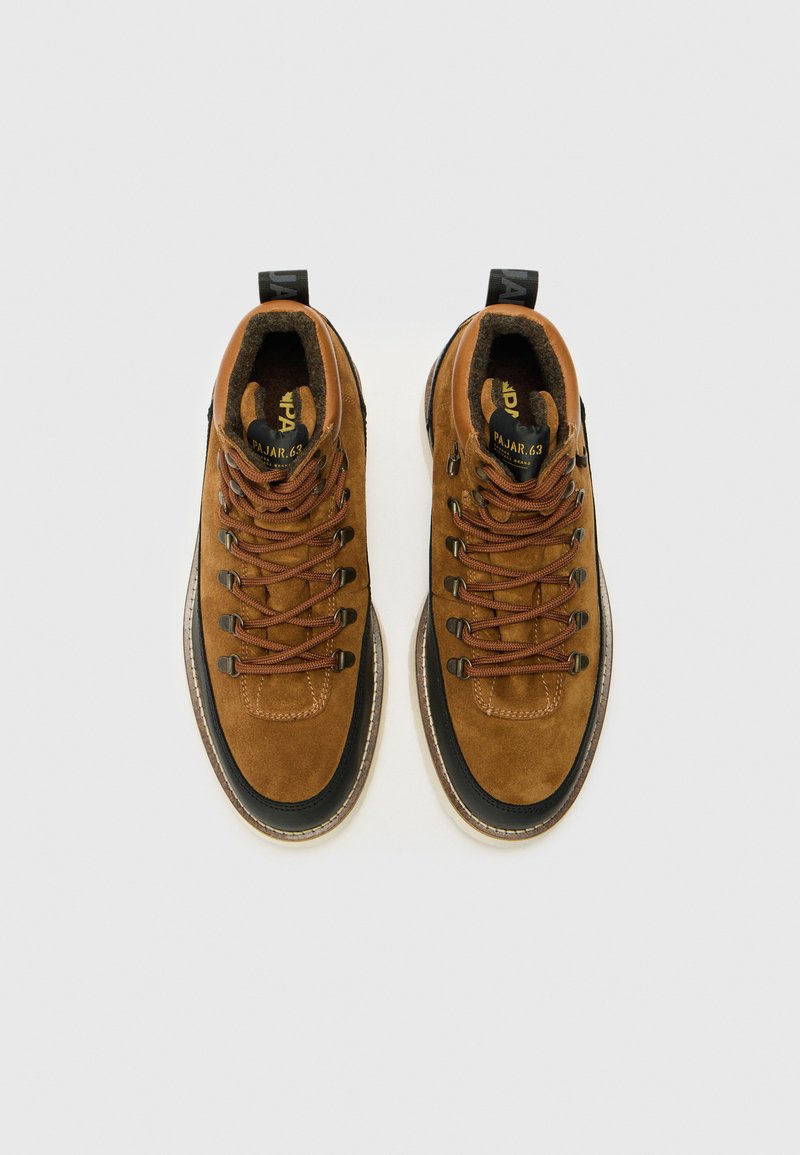 Bruine suède enkellaarsjes met zwarte rubberen zolen, voorzien van bruine veters, versterkte neus en een trekband voor gemakkelijk aan- en uittrekken. Vlakke kraag, textuuraccenten.