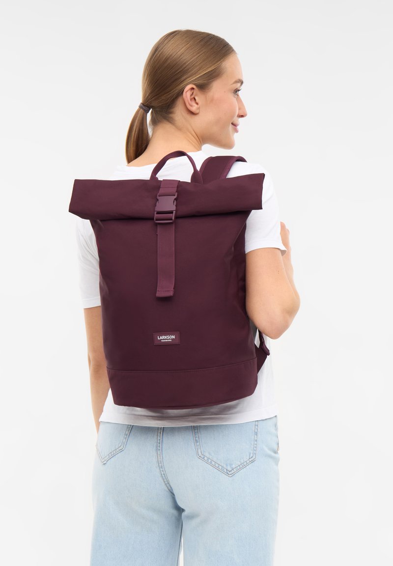 Femme aux cheveux châtain clair en queue de cheval se tient de côté, vêtue d'une chemise blanche, d'un jean bleu clair et d'un sac à dos violet foncé à rebord roulé.