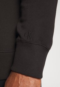 Czarna, długi rękaw bluzy z mankietem wykonanym z elastycznej tkaniny, o prążkowanej fakturze i małym, wytłoczonym logo "cK".