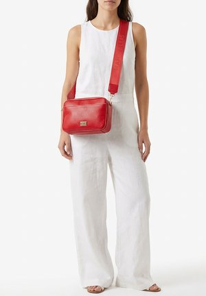 Vrouw draagt een witte mouwloze jumpsuit met wijde pijpen en draagt een rode leren crossbodytas met brede riem over de schouder.