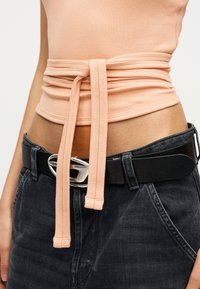 Γυναίκα που φοράει μαύρο τζιν με ψηλή μέση και μαύρη ζώνη, με τυλιγμένο ριμπ πουά κορσέ σε ροδακινί χρώμα δεμένο πάνω από ροδακινί μπλούζα.