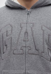 Gros plan d'un sweat à capuche gris zippé avec des lettres en relief formant le mot GAP sur la poitrine et une fermeture éclair visible au centre.