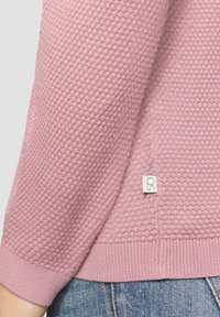 Gestructureerde roze trui met een honingraatpatroon, geribde manchetten en een subtiele logo-label op de zijkant gestikt. Klassiek ontwerp met ronde hals.
