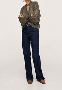 Blouse verte fluide à motif cachemire, col boutonné ; associée à un jean évasé bleu foncé et des escarpins marron à bouts pointus.