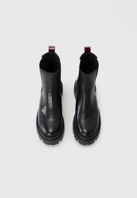 Bottines noires avec texture crocodile, bout rond, semelle épaisse et languette bordeaux. Fabriquées en cuir avec une finition mate.