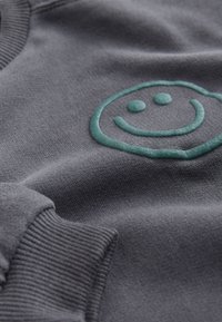 Tissu gris avec bord-côte et un motif visage souriant texturé bleu sarcelle à la surface.