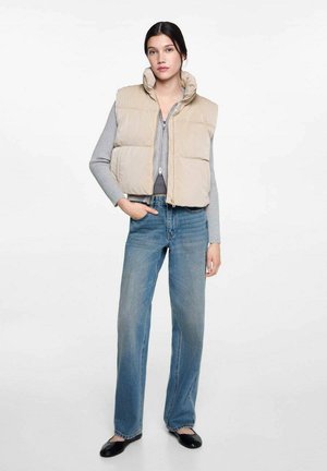 Gilet imbottito beige con colletto alto, chiusura con zip, indossato sopra una maglietta grigia. Abbinato a jeans larghi blu chiaro e ballerine nere.