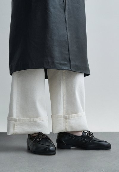 Svarta läderskor med dekorativa snören, kombinerade med vita vida jeans med uppvikta fållar, bärna under en svart läderrock.