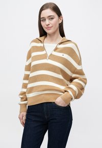 Bézs és fehér csíkos kötött melegítőfelső cipzáros gallérral és laza szabással, sötétkék farmerrel párosítva. Puha tapintású és laza ujjú.