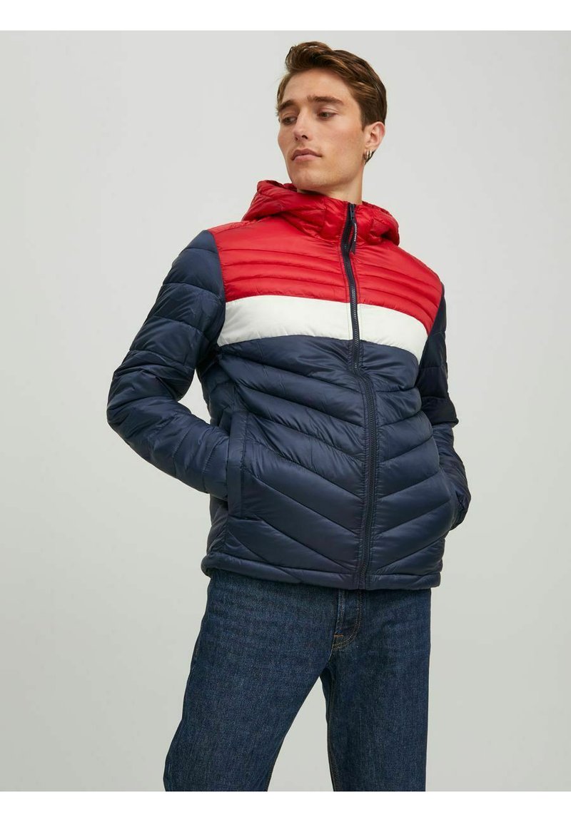 Padded bunda s kapucí, která má červený a bílý horizontální pruh přes hrudník, navy modrou barvu a zipový uzávěr.