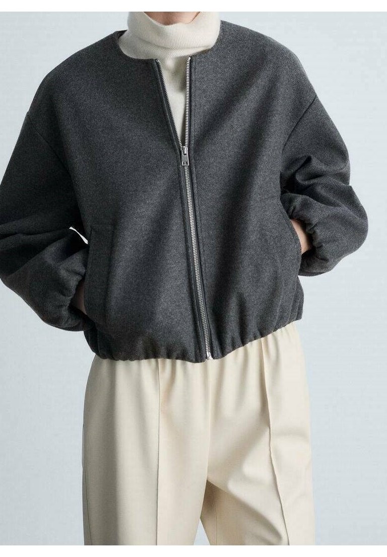 Grijze cropped jack gemaakt van zachte stof, met een rits aan de voorkant, elastische manchetten en een ronde halslijn. Het model draagt crèmekleurige broek.