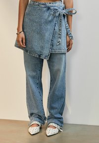Denim suknja obavijena oko tijela s remenom, složena preko svijetloplavih širokih traperica. Bijele cipele s oštrim nosom i zakovicama upotpunjuju izgled.