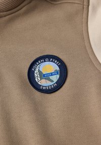 Beige stof med en cirkulær blå plet med et landskabsdesign, sol og tekst "POLARN O. PYRET SWEDEN SINCE 1976." Teksturiseret ribbet krave.