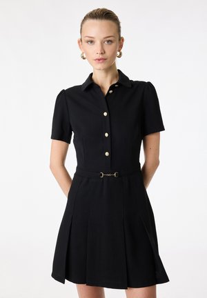 Vrouw draagt een zwarte korte mouwen jurk met gouden knopen aan de voorkant en een dunne riem in de taille, staande tegen een witte achtergrond.