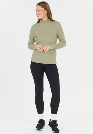 Junge Frau lächelnd, trägt ein hellgrünes langärmliges Oberteil mit Reißverschluss, schwarze Leggings, weiße Socken und schwarze Laufschuhe, steht vor weißem Hintergrund.