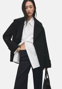 Zwarte oversized blazer met een dubbelgeknoopte sluiting, gecombineerd met een wit overhemd en marineblauwe broek met krijtstreepjes. Zwarte draagtas.