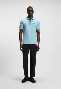 Ljust blå polo-tröja med svarta och vita ränder längs kragen, kombinerad med svarta byxor och svarta skor. Enkel, klassisk design.