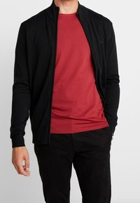 Czarna kurtka na zamek z prążkowanym kołnierzem i mankietami, nałożona na jednolity czerwony T-shirt. Gładka tkanina, dopasowany krój i minimalne zdobienia.