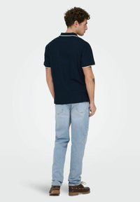 Man die wegkijkt en een marineblauw poloshirt met witte accenten draagt, lichtblauwe spijkerbroek en donkerbruine veterschoenen tegen een effen achtergrond.