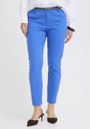 Femme portant un pantalon bleu vif ajusté avec une ceinture marron, une chemise blanche, un bracelet noir et des chaussures à talons marron avec bride arrière.