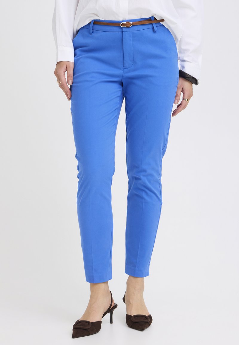 Žena oblečená v jasne modrých úzkych nohaviciach s hnedým opaskom, bielou košeľou, čiernym náramkom a hnedými sandálmi na podpätku so zadným remienkom.