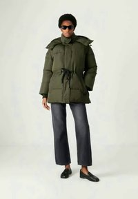 Ελιάς πράσινη φουσκωτή ζακέτα με ρυθμιζόμενη μέση, ψηλό γιακά και μεγάλη κουκούλα, συνδυασμένη με φαρδιά τζιν παντελόνια από σκούρο τζιν και μαύρες λοφάδες.