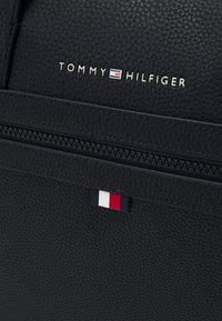 Sac à dos en cuir noir texturé avec une poche avant zippée, arborant un petit emblème rouge, blanc et bleu ainsi qu'un logo de marque argenté.