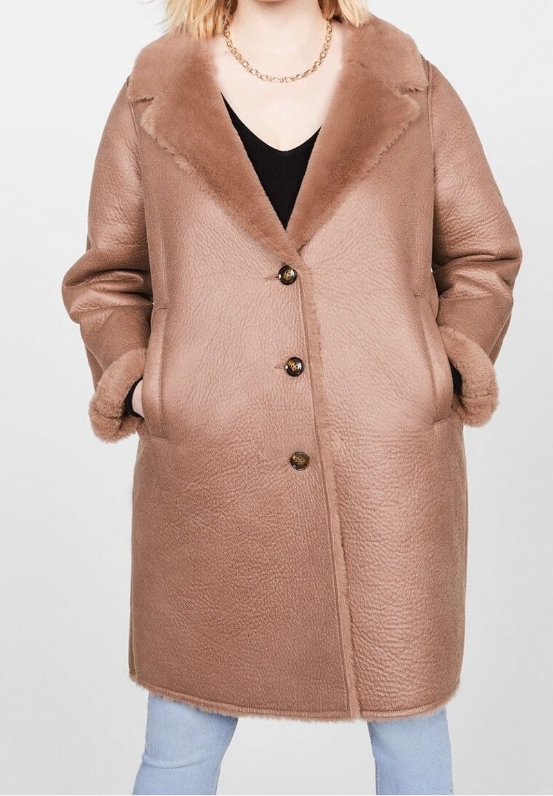 Femme portant un manteau en peau retournée couleur camel mi-long, avec trois boutons, des manches retroussées et un large col, sur un jean bleu clair.