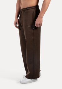 Mann trägt braune, weit geschnittene Sweatpants mit seitlichen Taschen und weiße Sneaker, steht vor einem schlichten weißen Hintergrund.