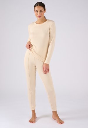 Vrouw staat blootsvoets, gekleed in een beige top met lange mouwen en bijpassende nauwsluitende broek, tegen een effen lichte achtergrond.