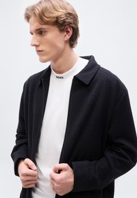 Μαύρο υφασμάτινο μπουφάν με γιακά πάνω από ένα λευκό μπλουζάκι με στρογγυλή λαιμόκοψη που φέρει λογότυπο. Σιλουέτα στενής εφαρμογής και κλείσιμο με φερμουάρ. Χέρια σταυρωμένα στο στήθος.