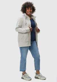 Veste zippée gris clair avec un motif moucheté, dotée d'un col haut et de poignets côtelés, associée à un jean bleu et des baskets grises.