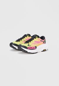 Coppia di scarpe da corsa Hoka in giallo neon, rosa e nero con suole spesse bianche e lacci neri su sfondo bianco.