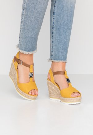 Żółte sandały na koturnie z plecioną jutasową podeszwą, z brązowymi paskami wokół kostki i dekoracyjnym akcentem z logo w pobliżu palców.