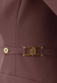 Tessuto mauve con cuciture sartoriali, un accento di fibbia in tono oro con un design geometrico e un dettaglio di bottone dorato.