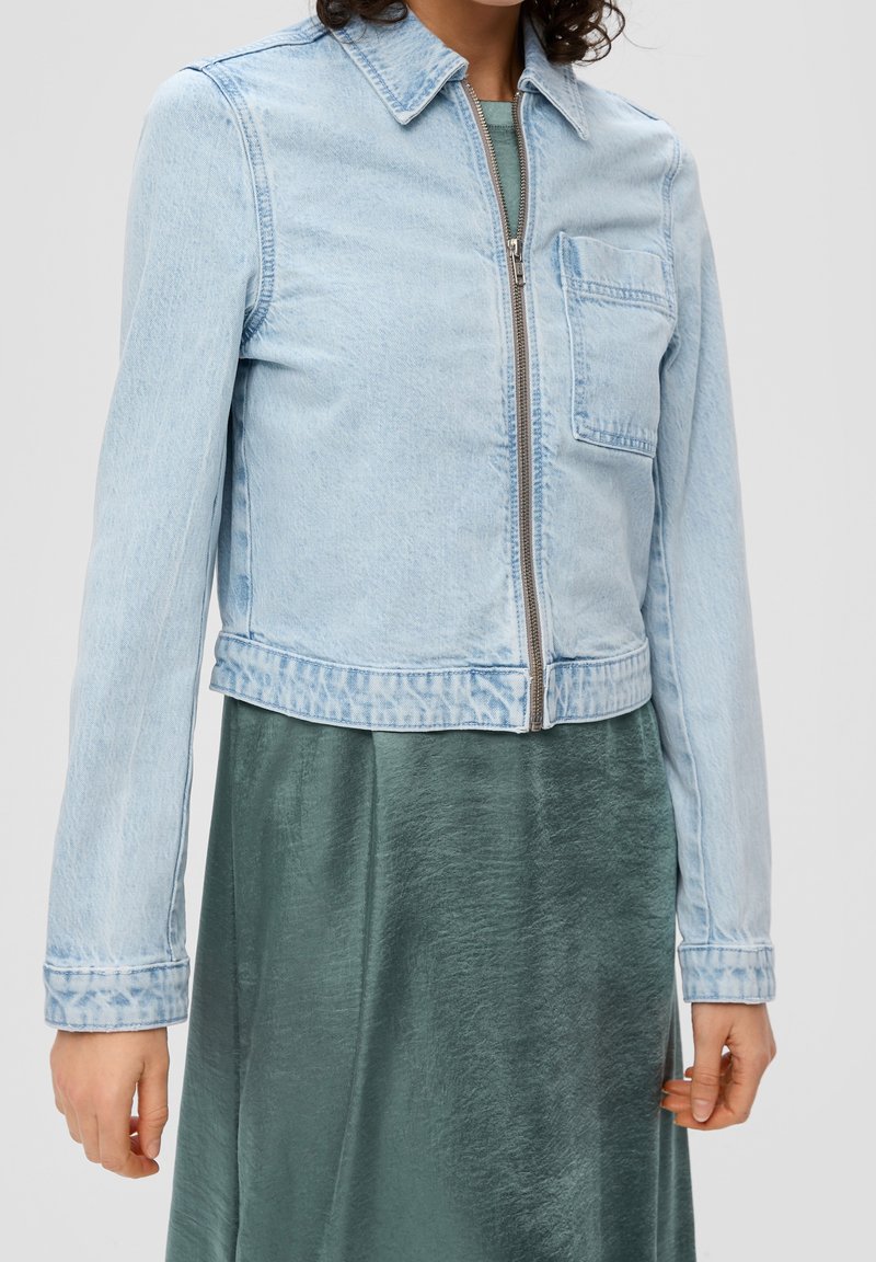 Helle blaue Jeansjacke mit Reißverschluss, Kragen und Vordertasche. Hat ein cropped Design und einen elastischen Saum. Trägt man über einem grünen Kleid.