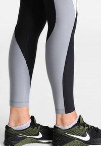 Ben klädda i svarta och grå träningsleggings och svarta och gröna Nike löparskor med vita swoosh-logotyper mot en vit bakgrund.