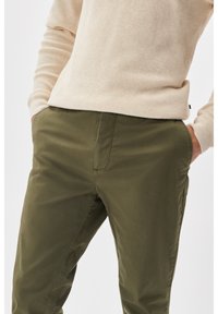 Olive green cotton pants with flat front, side pockets, and a smooth texture, paired with a light beige knitted sweater.
