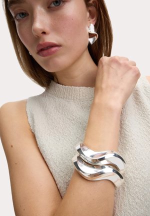 Sculpturale zilveren oorbellen en een cuff armband hebben een glad, golvend ontwerp. Draag ze bij een textuurrijke crèmekleurige top, zodat de sieraden het licht weerkaatsen.