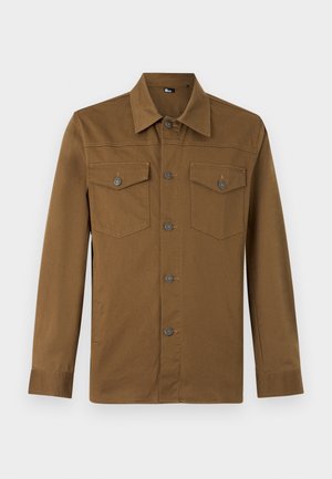 Mann iført en brun jakke med knapper over en hvit skjorte, blå jeans og hvite joggesko, står mot en ensfarget lys vegg.