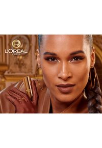 Rossetto L'Oréal Paris in un tubo dorato tenuto da una persona che indossa un guanto di raso marrone, con una giacca di pelle marrone e grandi orecchini a cerchio.