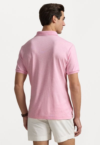 Camisa polo rosa de manga corta con cuello clásico, con una textura lisa y detalles de abertura lateral. Combinada con pantalones cortos de color claro.