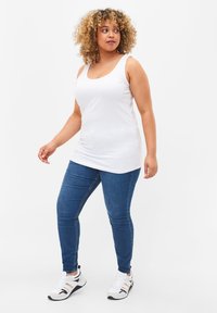 Biały top na ramiączkach z okrągłym dekoltem, w połączeniu z niebieskimi slim jeansami. Model nosi białe trampki z czarnymi i złotymi akcentami. Prosty design.
