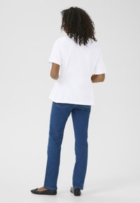 Witte kortemouwen hemd met een cinched taille, gecombineerd met blauwe recht gesneden jeans en zwarte platte schoenen, gezien van achteren.