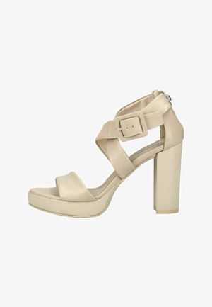 Sandale beige à talon haut avec larges lanières croisées, grande fermeture à boucle et talon bloc épais sur fond blanc.