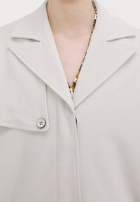Lysegrå blazer med en stor krave, knaplukninger og et struktureret stof. Underneath er en mønstret bluse delvist synlig.