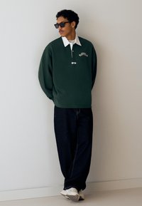 Grøn pullover med hvid krave, broderet detalje, afslappet pasform, kombineret med mørke jeans og hvide sneakers. Glat stof og enkel design.