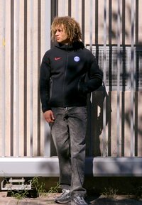 Sort zip-up jakke med rødt Nike-logo, blåt rundt mærke; grå jeans; står op ad en lodret panelbaggrund.