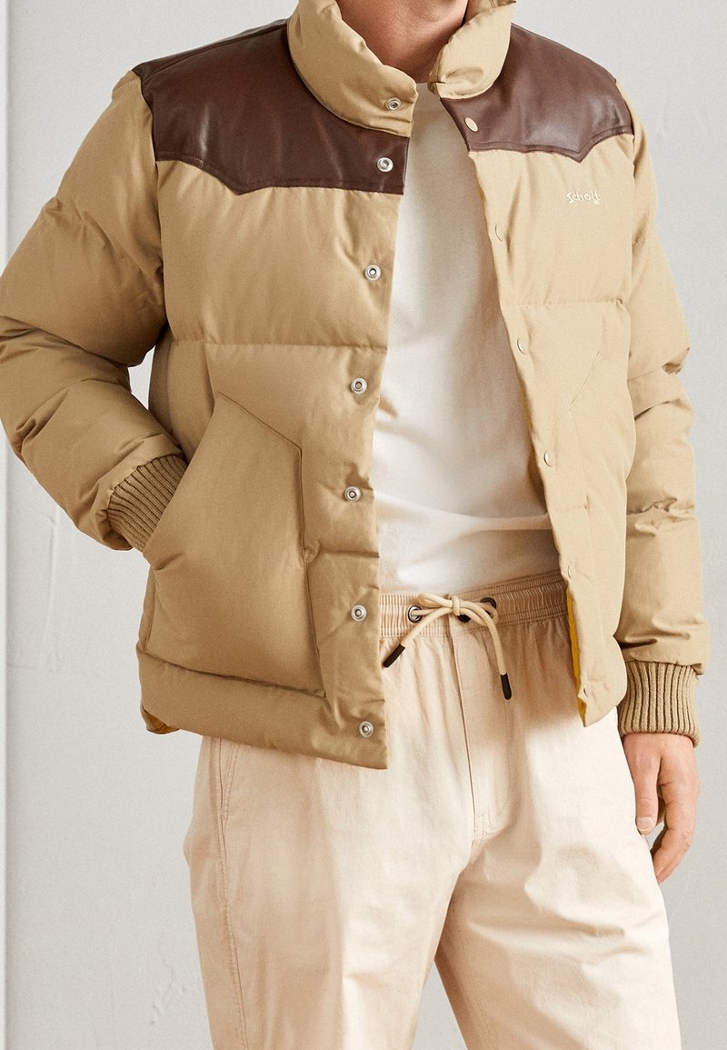 Homme portant une doudoune beige avec des panneaux d'épaule marron, une chemise blanche et un pantalon beige clair à cordon, debout avec une main dans la poche.